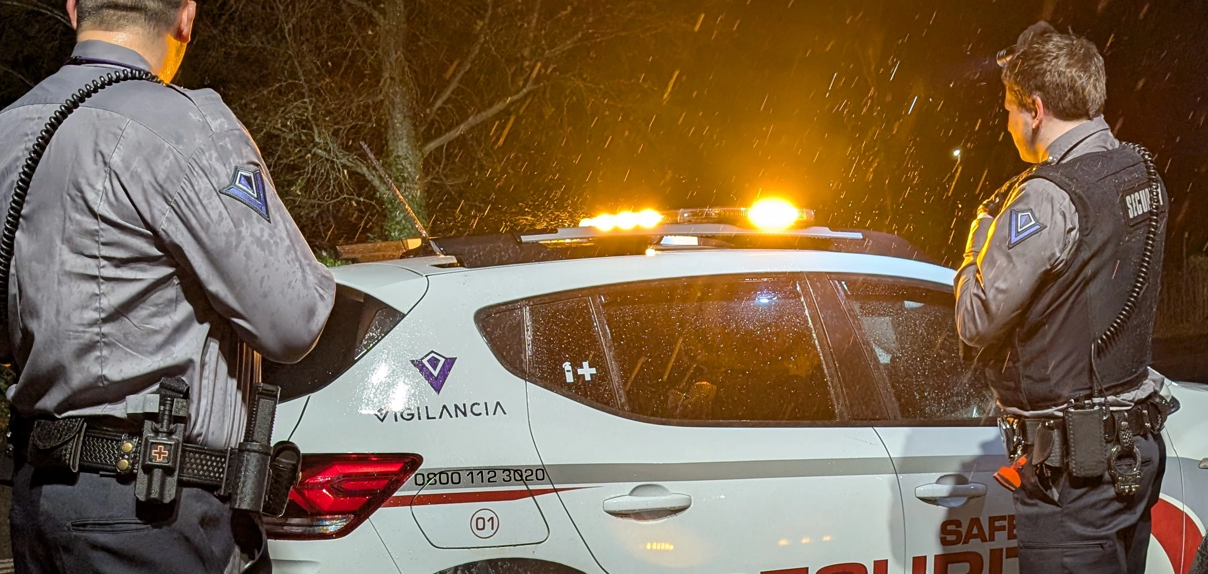 Two security guards standing in front of a patrol car with amber lights flashing.